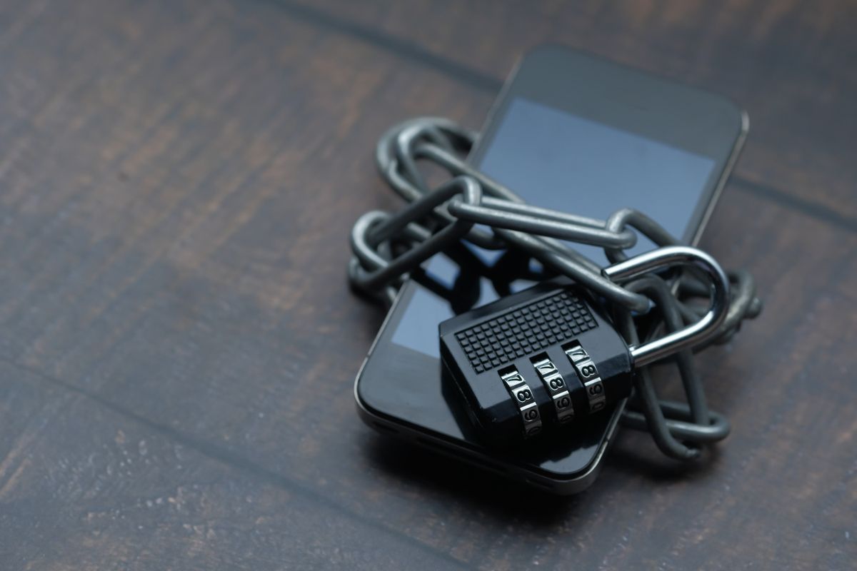 A modern smartphone wrapped in a metal chain and secured with a combination padlock on a wooden desk, symbolizing locking down phone data before a reset
