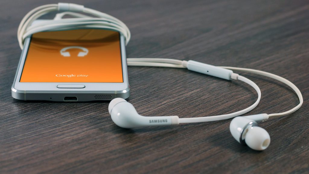 A smartphone showing a music screen beside a pair of white earbuds on a dark wooden desk, illustrating a Bluetooth audio connection that keeps disconnecting