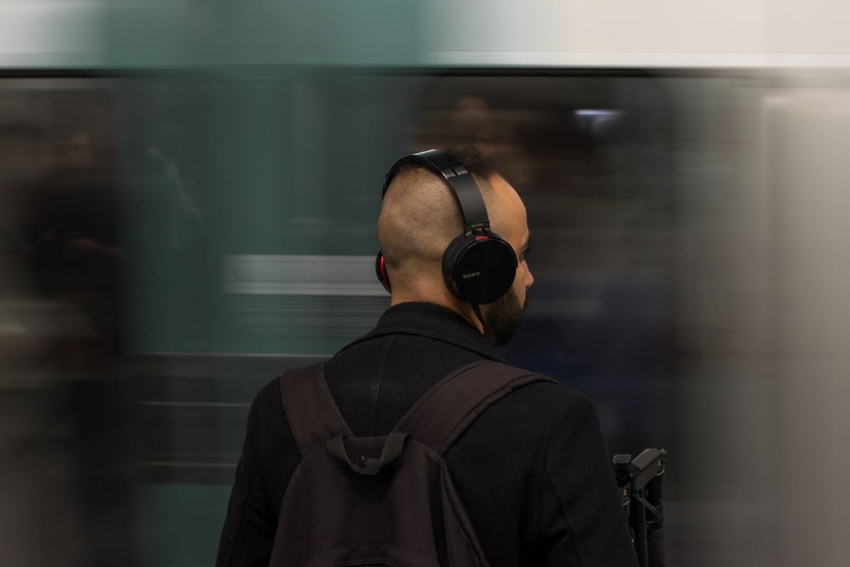 Commuter on a subway platform wearing black over-ear headphones as a train blurs past behind him