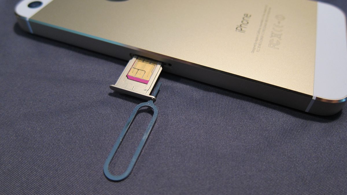 A gold iPhone 5S with its SIM tray ejected and a nano-SIM sitting in it, the SIM-eject pin resting below, showing how a physical SIM pops out to move between phones
