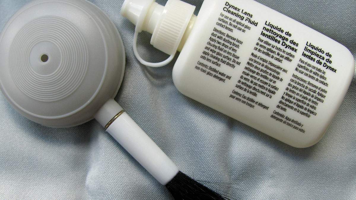 A lens cleaning fluid bottle with its printed instruction label facing the camera, next to a brush and cloth