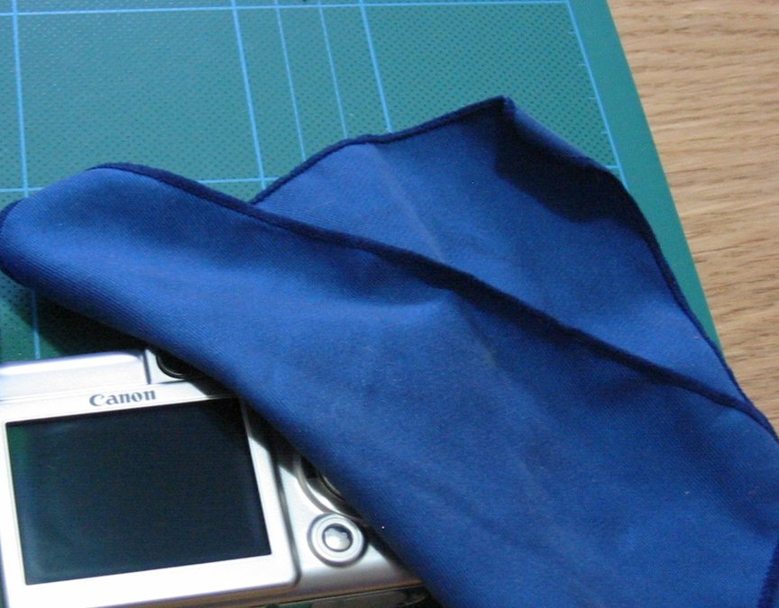 A blue microfiber cloth draped over a switched-off compact camera screen on a green cutting mat