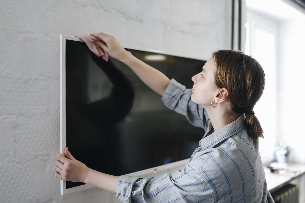 A person wiping a wall-mounted flat screen with one hand while the other hand steadies the panel by its edge
