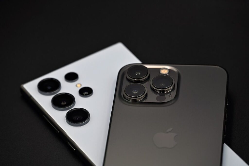 A graphite iPhone 13 Pro and a Samsung Galaxy S24 Ultra Android phone lying face-down side by side on a black surface with their cameras facing up, illustrating the iPhone vs Android choice