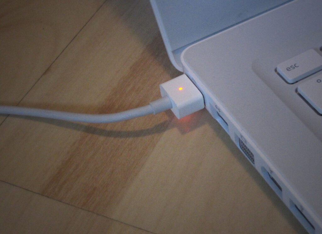 A white MacBook plugged into a MagSafe charger with the connector's charge light glowing amber