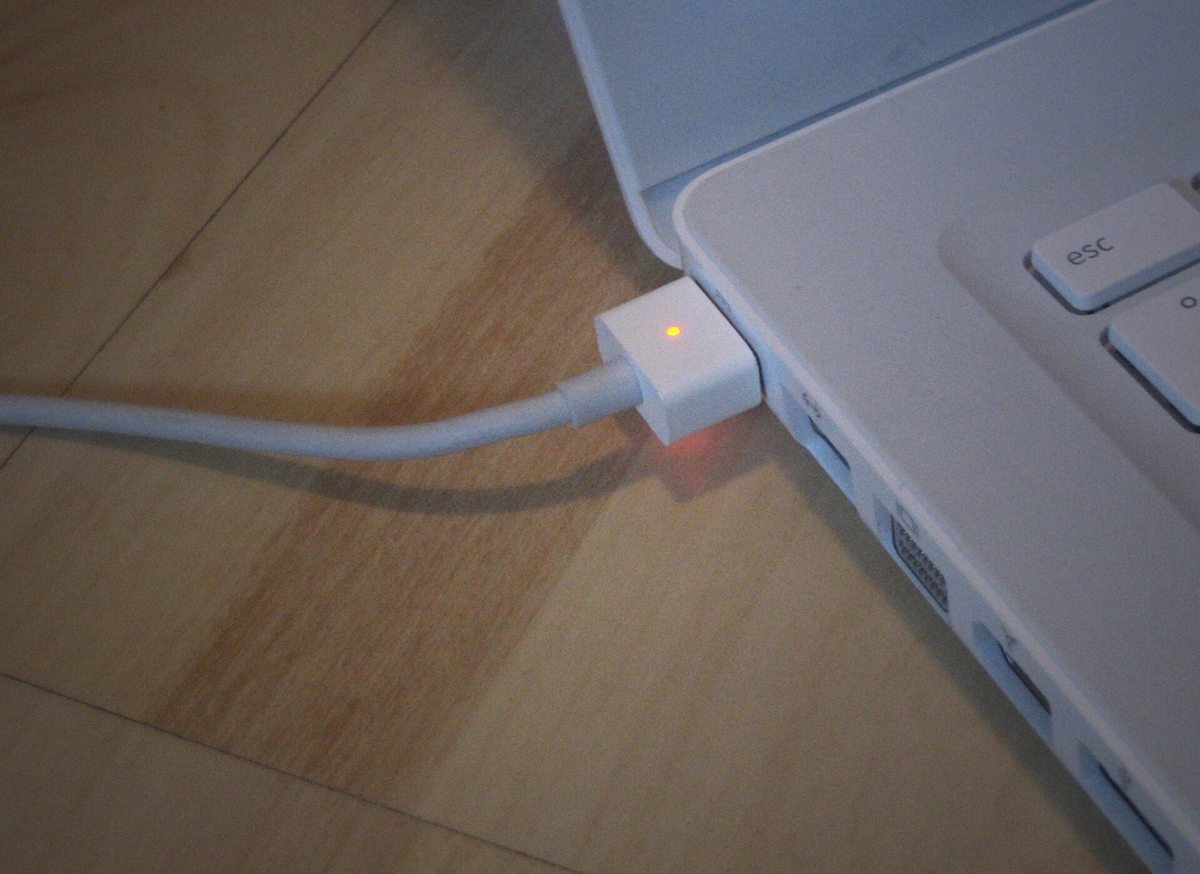 A white MacBook plugged into a MagSafe charger with the connector's charge light glowing amber
