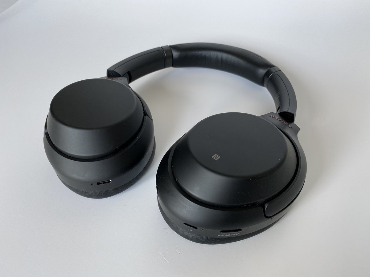 A pair of black Sony WH-1000XM3 over-ear headphones on a white surface, the power and NC/AMBIENT control buttons visible on the bottom edge of the earcup