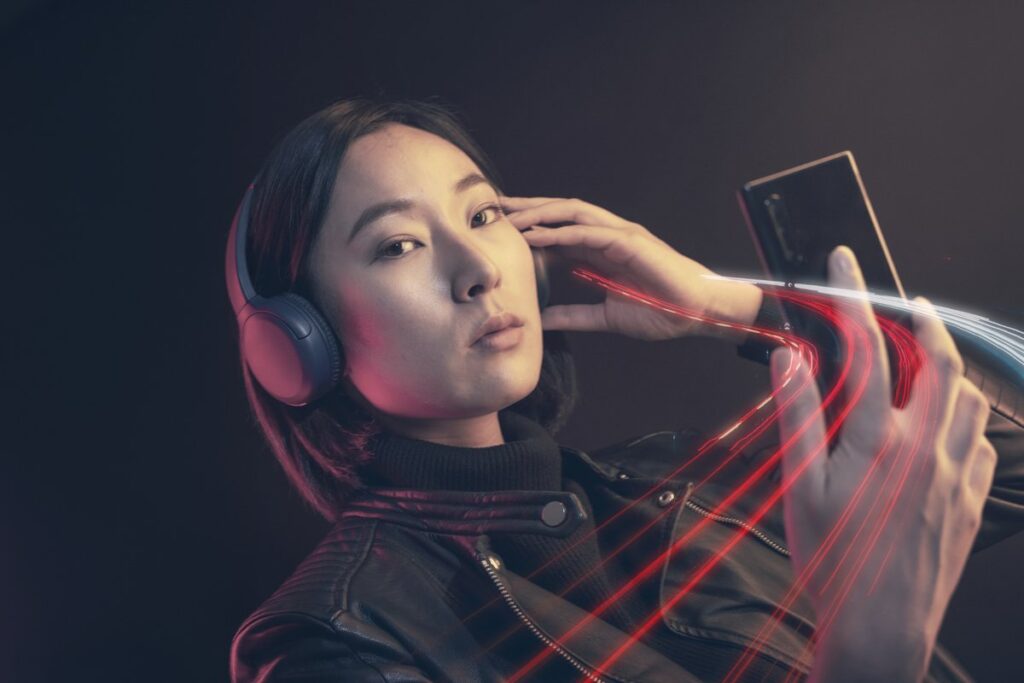 A woman wearing black over-ear Bluetooth headphones looks at her smartphone, red light trails streaming from the phone to the headphones to suggest a wireless connection