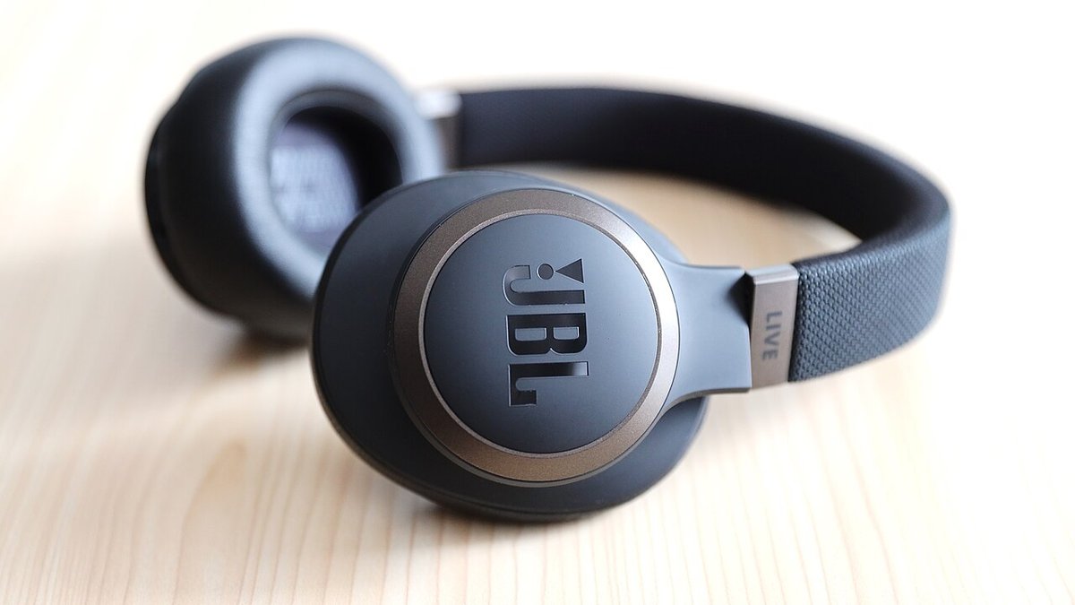 Black JBL LIVE 650BTNC over-ear Bluetooth headphones lying on a wooden desk, JBL logo earcup facing the camera