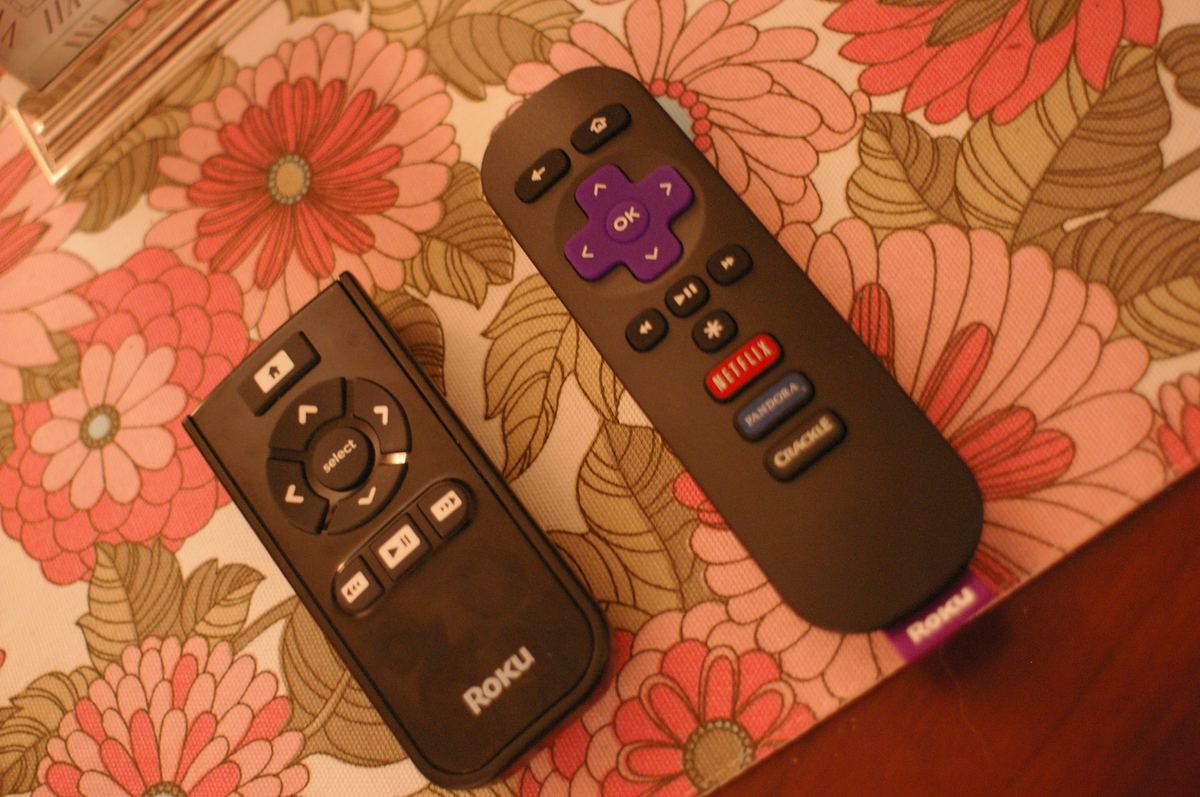Two real Roku remotes side by side on a floral cloth: a simple IR remote on the left with just a D-pad and playback keys, and an enhanced remote on the right with Netflix, Pandora and Crackle shortcut buttons