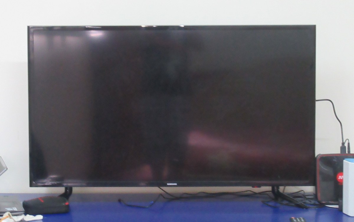A real Samsung LED TV on a desk showing a completely black screen with only a faint reflection, the Samsung logo visible on the bottom bezel