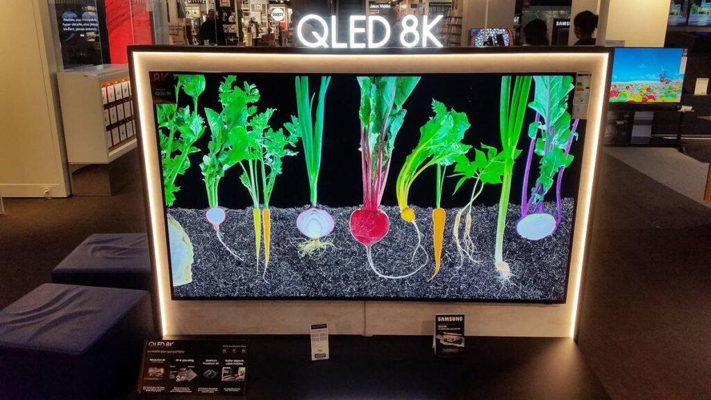 A large QLED television on a store wall showing vivid vegetables against a black background, the kind of bright saturated color an RGB LED TV aims to deliver