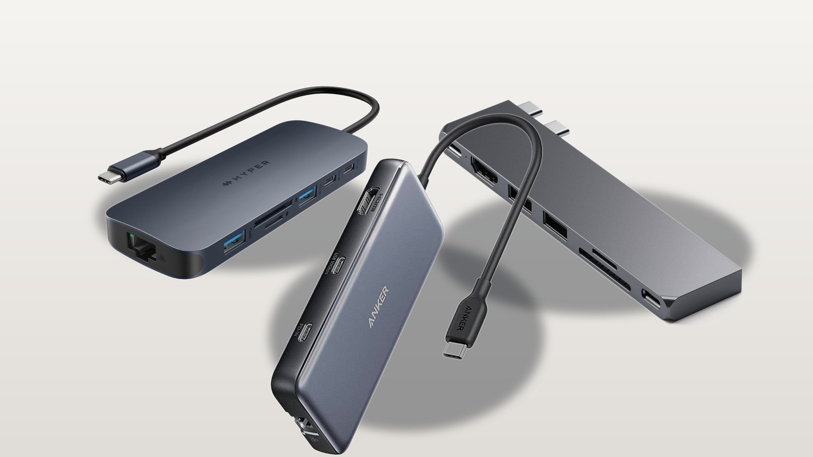 Three real USB-C hubs for MacBook, a HyperDrive Next, an Anker 555, and a Satechi Pro Hub Slim, arranged on a neutral studio background